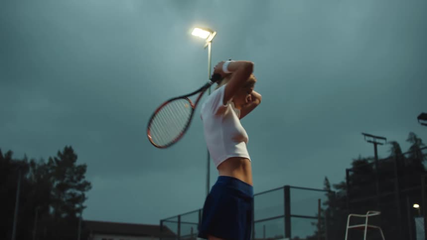 A determined female tennis player practices her serve in slow motion on an outdoor court under dramatic evening lights