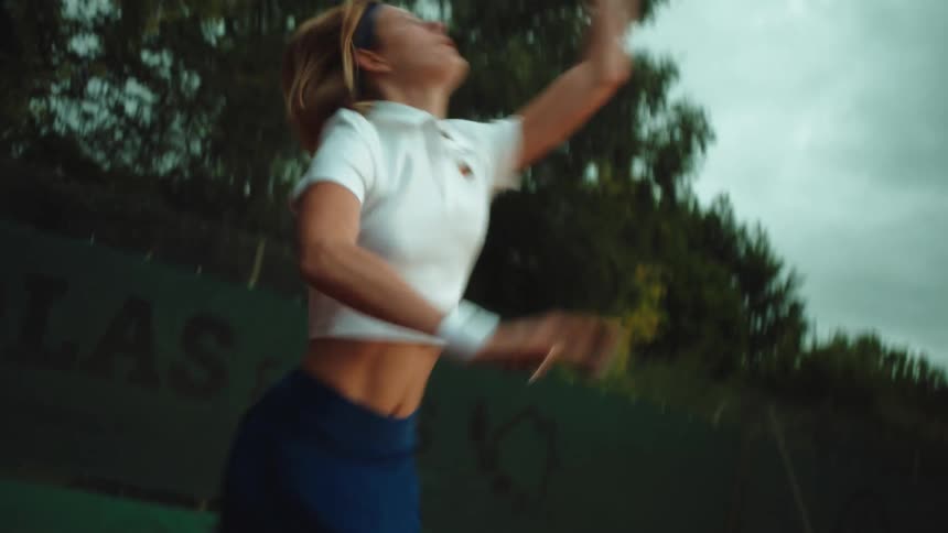 A focused female athlete in a vintage-style outfit serves a tennis ball in slow motion on an outdoor clay court