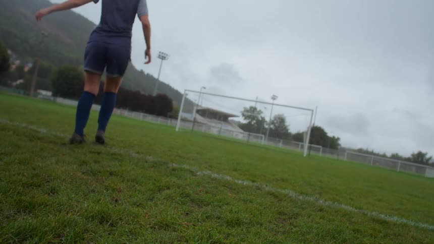 From a low angle, a female soccer player kicks a ball towards the goal in slow motion on a cloudy day
