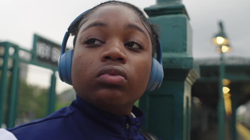 A close-up of a young woman wearing headphones with a thoughtful expression while waiting at an elevated train station