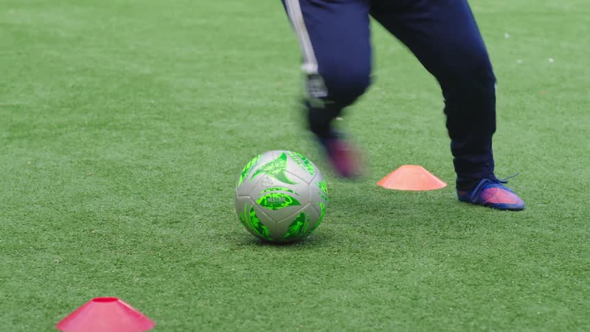 A close-up shot shows a soccer player's feet skillfully dribbling a ball around training cones on a turf field