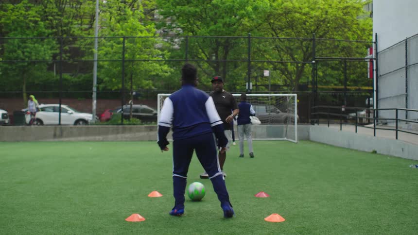 From behind, a young athlete in a blue tracksuit practices soccer dribbling skills as their coach watches on a green turf field