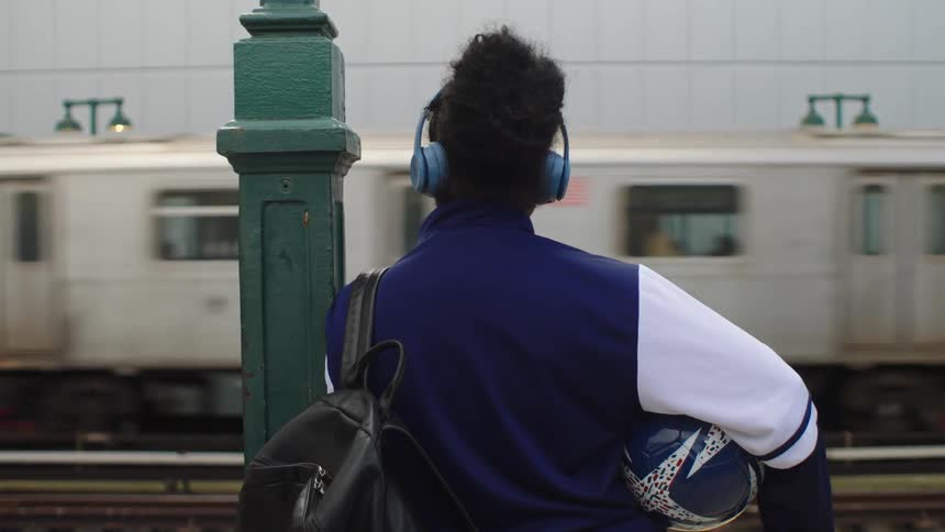 A young athlete with headphones and a soccer ball waits on a subway platform as a train passes by