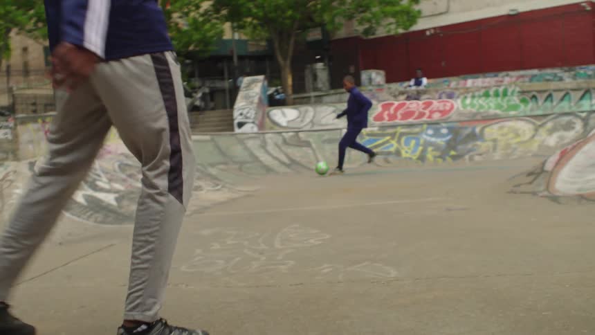 Two friends practice their soccer skills in slow motion at a graffiti-covered urban skatepark