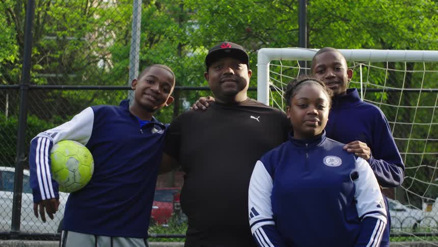 A proud African American father smiles for a portrait with his three athletic children on a sunny soccer field