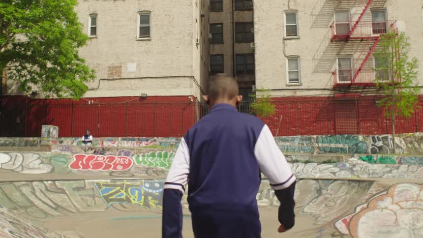 Two young boys in tracksuits practice soccer skills in slow motion, passing a ball back and forth inside a graffiti-covered urban skatepark