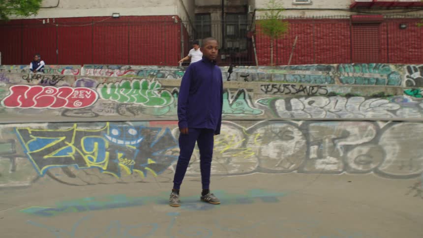 A young boy in a purple tracksuit practices soccer in a graffiti-covered skate park and then stands looking thoughtfully into the distance