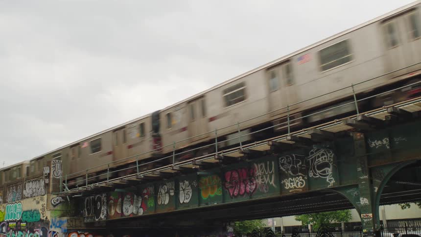 An urban subway train passes over a graffitied elevated track on an overcast day in the city