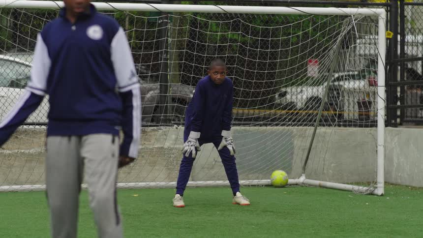 A young boy in a goalie uniform makes a spectacular save in slow motion during soccer practice on an urban field