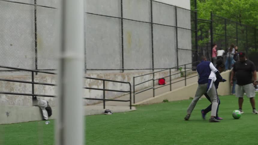 A young soccer player dribbles past a defender and kicks the ball into the goal while a coach watches on at an urban park