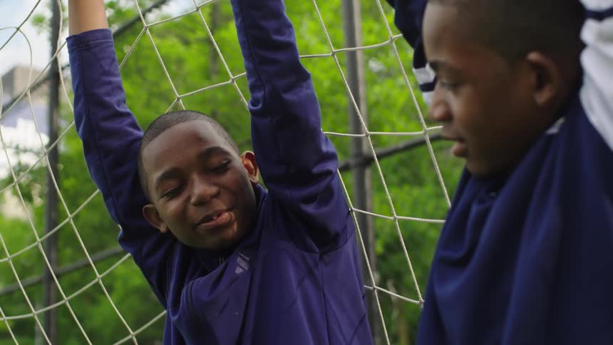 Two young African American boys in soccer uniforms laugh and talk together while one hangs playfully on the goal net