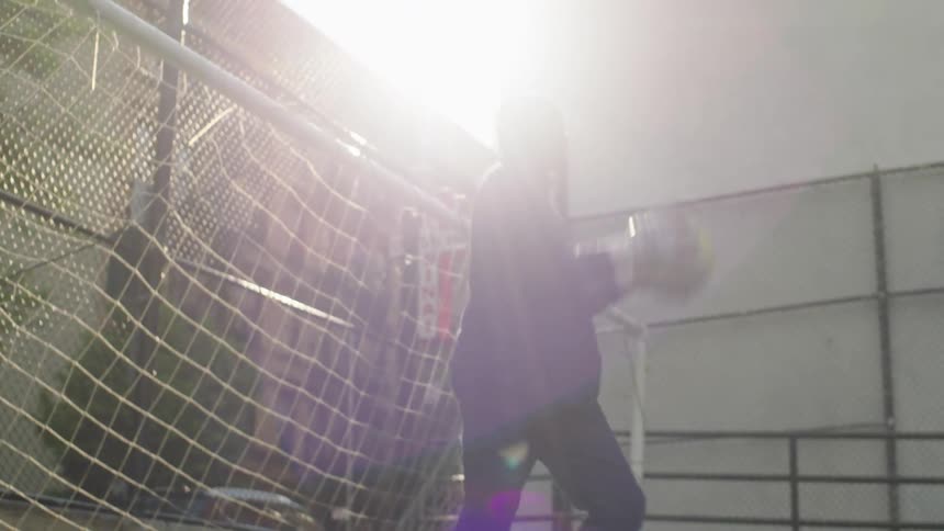 A young boy practices his goalkeeping skills with a soccer ball on a sunny urban turf field in slow motion