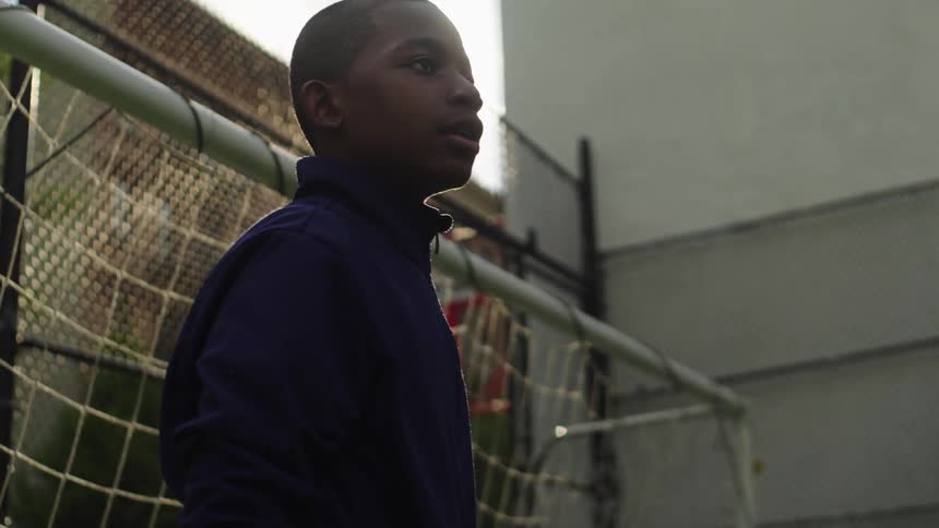 A young Black boy in a blue sweatshirt acts as a soccer goalie, catching the ball in slow motion as the sun flares behind him