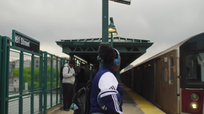 A person with headphones and a soccer ball waits on a subway platform as the train slowly pulls into the station on an overcast day