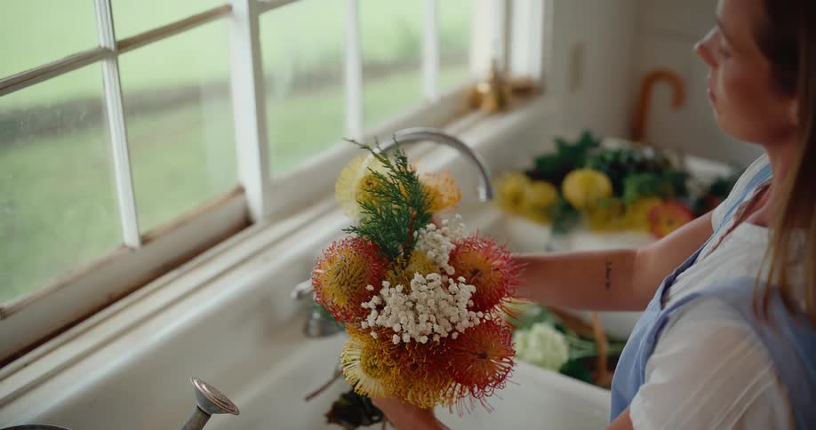 A woman carefully arranges a beautiful bouquet of colorful pincushion protea flowers in a rustic farmhouse sink
