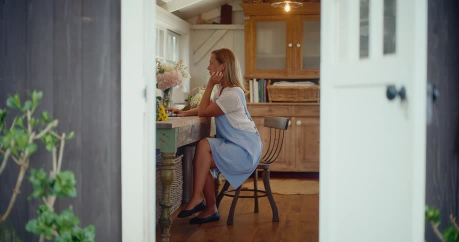 A woman works on her laptop at a rustic desk in a charming home office, seen through an open doorway