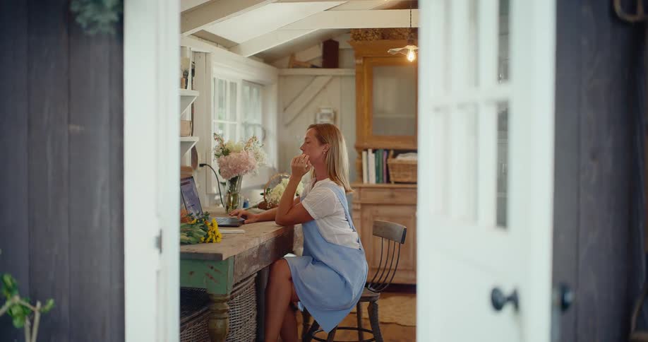 A woman works on her laptop in a cozy, rustic home office, viewed through an open doorway