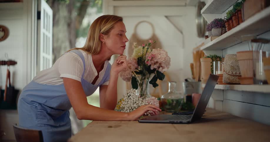Focused woman wearing an apron browses on her laptop inside her charming flower workshop