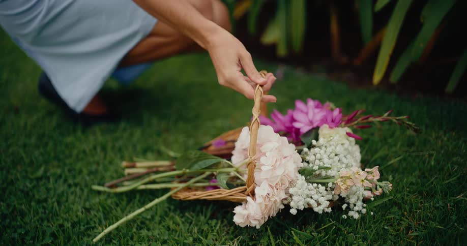 A woman in a blue dress gently places her wicker basket full of fresh flowers on the lush green grass