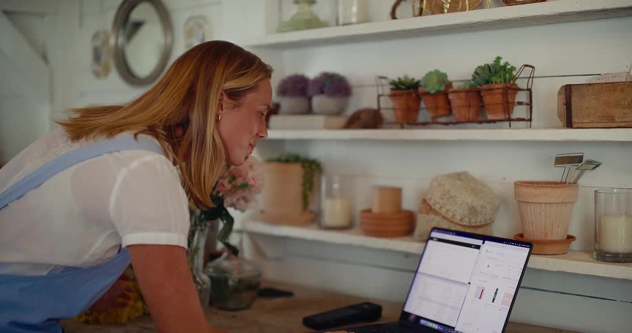 A smiling female florist arranges a beautiful bouquet in her workshop before getting stressed out while managing her business on her laptop