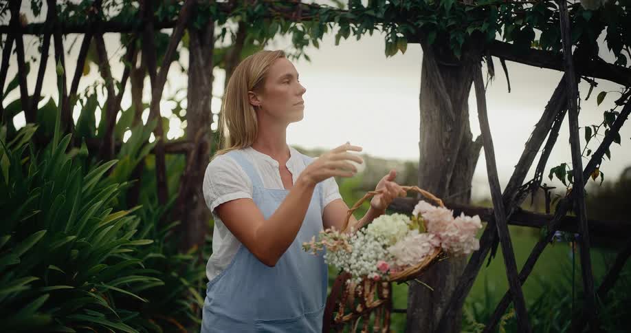 A serene woman in an apron holds a basket of fresh flowers and admires the beauty of her lush country garden at dusk