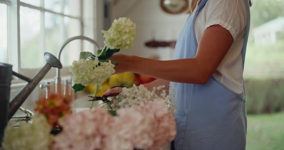 A woman in a blue apron stands at a rustic farmhouse sink, carefully trimming fresh white hydrangea flowers for a bouquet