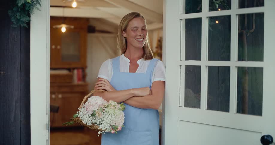 A smiling woman in an apron proudly holds a basket of fresh flowers while standing in the doorway of her rustic home