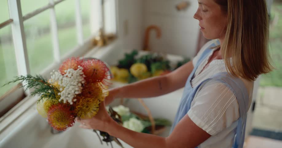 Young woman in a light-filled farmhouse kitchen carefully arranges a colorful and unique flower bouquet over the sink