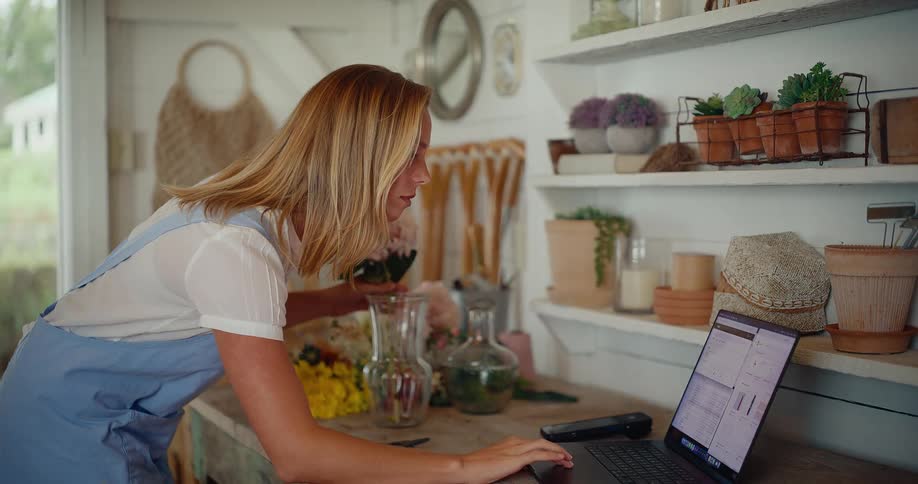 Young female florist manages her small business on a laptop while arranging a beautiful flower bouquet in her workshop