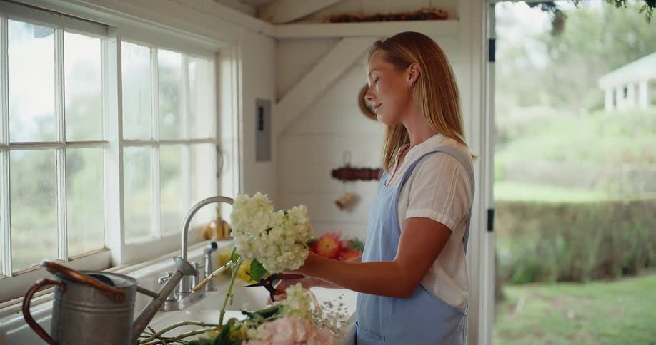 Woman in a blue apron mindfully arranging a fresh bouquet of hydrangeas in a rustic farmhouse kitchen by a window