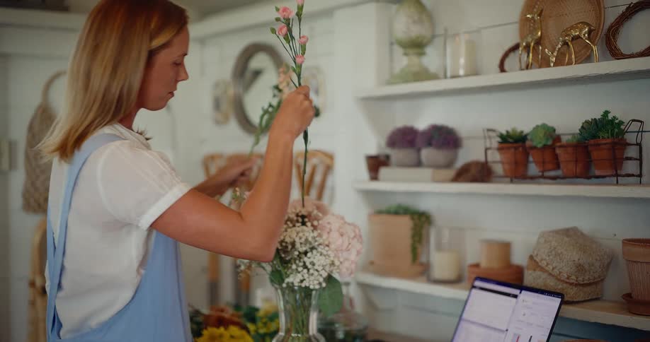 A female florist carefully arranges a beautiful bouquet of flowers in a vase inside her bright and rustic flower shop