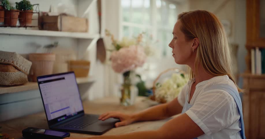 A female entrepreneur processes a credit card payment with a mobile reader and laptop in her rustic flower shop