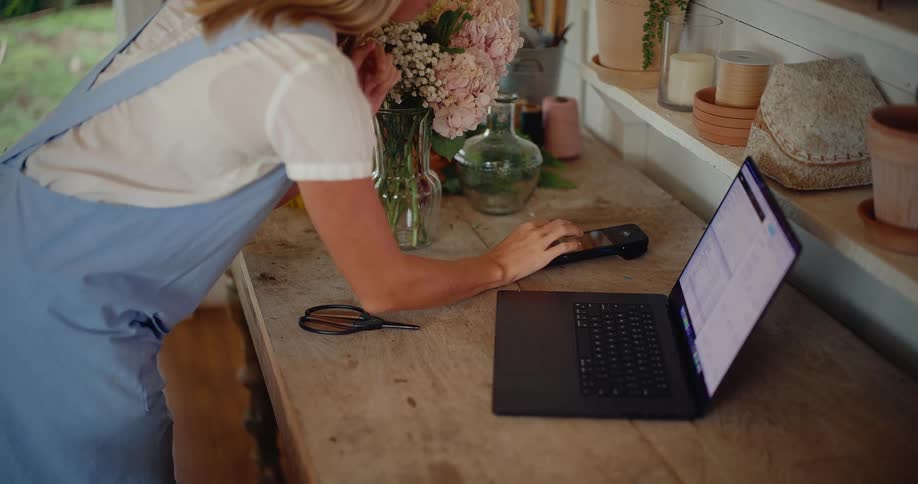 A female florist processes a payment and manages her business on a laptop in her charming flower shop