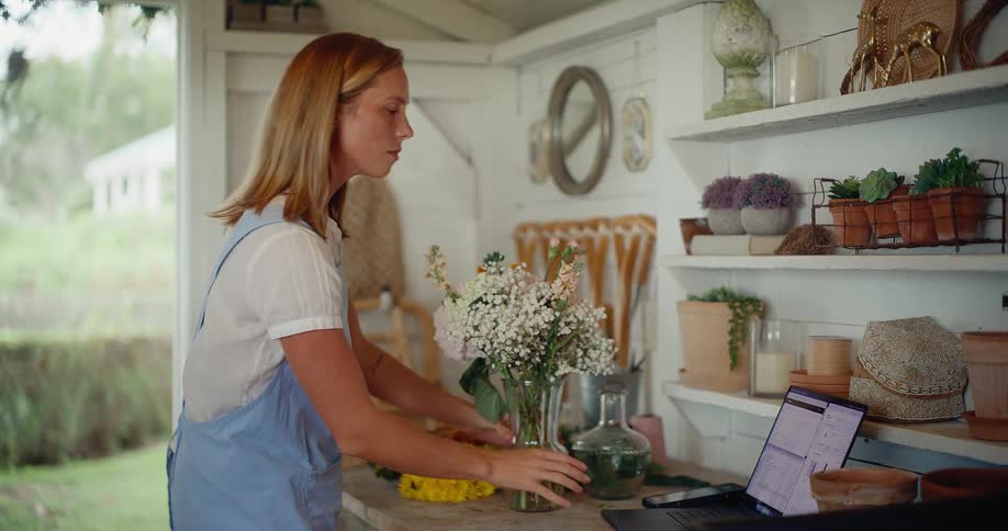 Young female florist arranges a beautiful flower bouquet in her charming workshop and manages her small business on her laptop