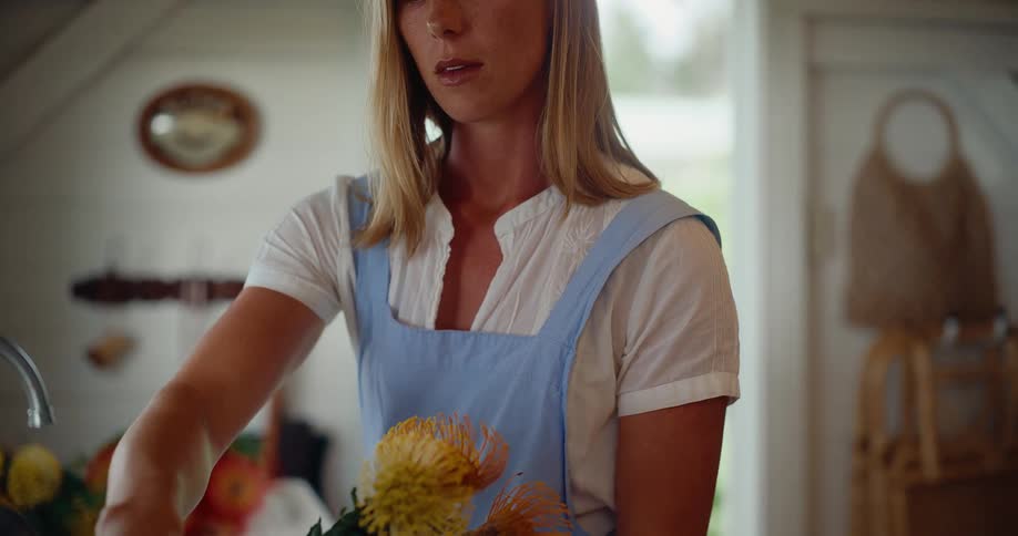 A woman in a blue apron mindfully arranges a beautiful bouquet of colorful flowers in her rustic kitchen