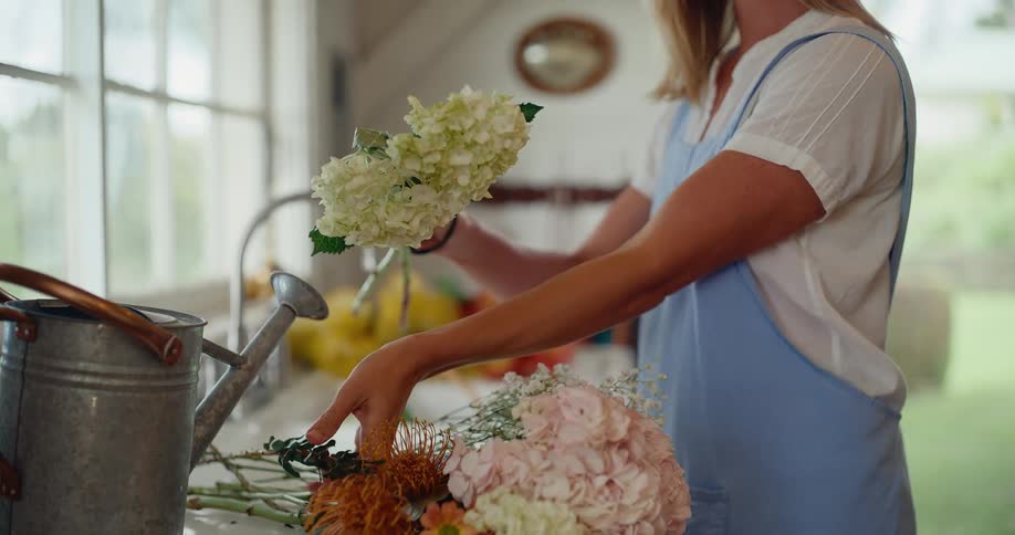 A woman in a blue apron carefully trims and arranges a beautiful bouquet of fresh flowers in her rustic kitchen in slow motion