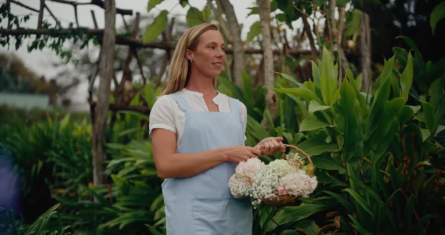 A beautiful woman in an apron walks in slow motion through a lush green garden, holding a basket of flowers