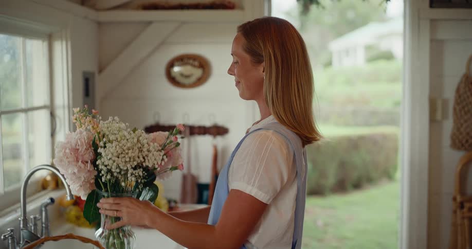 A florist in a blue apron carefully arranges a beautiful bouquet of pink hydrangeas in a glass vase inside her charming, rustic workshop