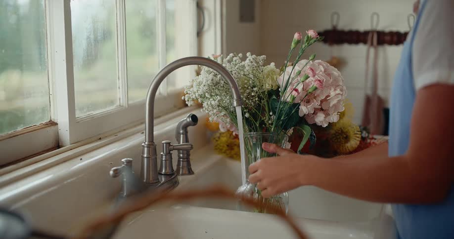 A woman's hands carefully fill a glass vase containing a beautiful bouquet of flowers with water from a rustic farmhouse kitchen sink