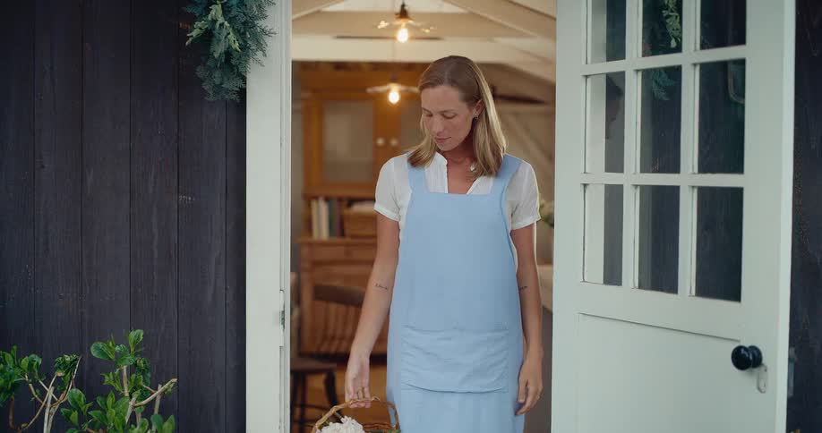 A charming woman in a blue apron holding a basket of fresh flowers stands in the doorway of her rustic home, smiling at the camera in slow motion