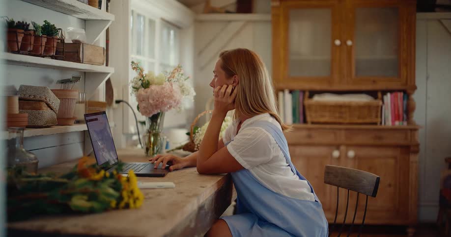 A thoughtful young woman works on her laptop computer in her beautiful and rustic flower workshop