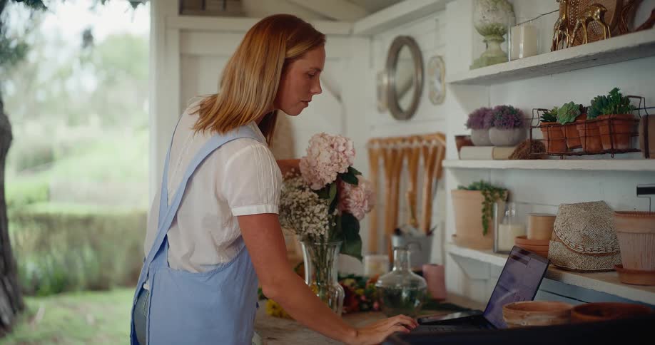 A young female florist in her charming workshop arranges a beautiful flower bouquet while managing her small business on a laptop