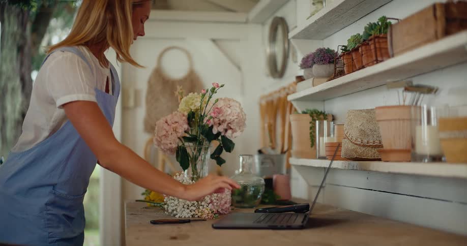 A female florist works on her laptop in her charming workshop before creating a beautiful flower arrangement