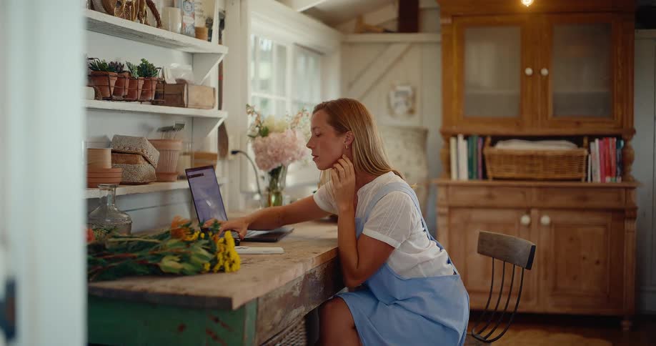 A florist works on her laptop computer in her rustic flower shop, managing her small business