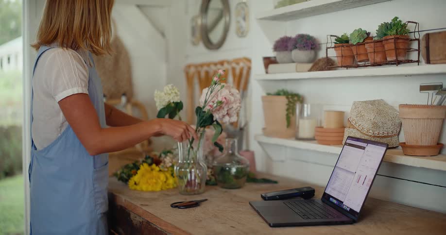 A female florist in an apron carefully arranges a beautiful bouquet of fresh flowers in a vase inside her rustic and charming workshop