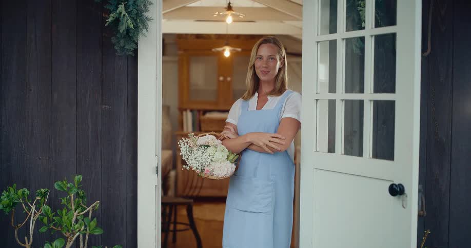 A smiling woman in a blue apron holding a basket of fresh flowers stands in the doorway of her cozy home