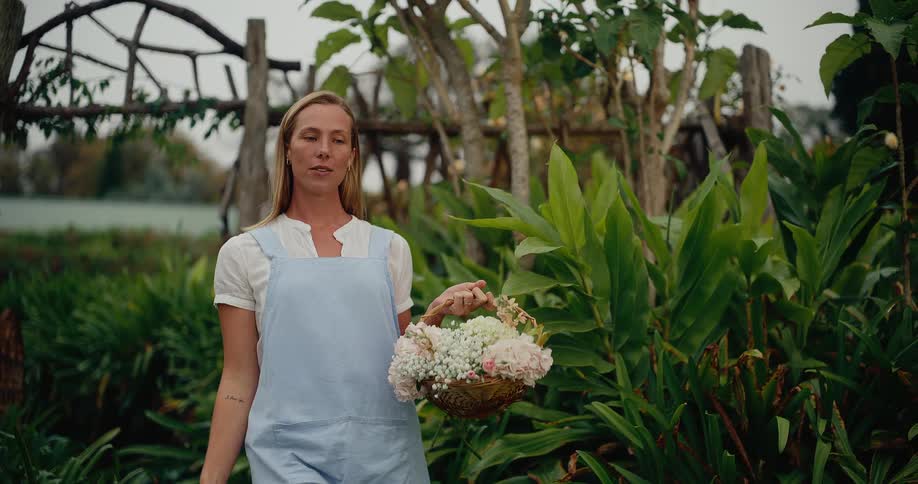A beautiful woman in an apron walks through a lush green garden in slow motion, holding a basket of freshly picked flowers