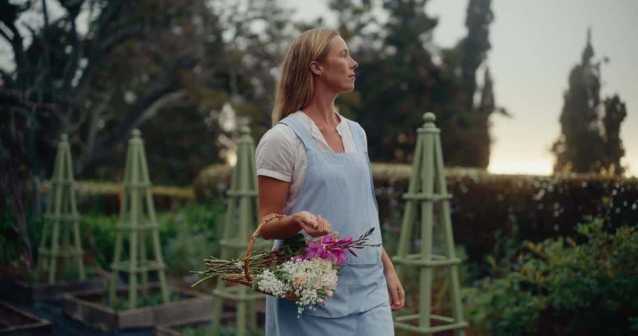 A tranquil female gardener in an apron strolls through her beautiful garden at dusk, holding a basket of freshly cut flowers