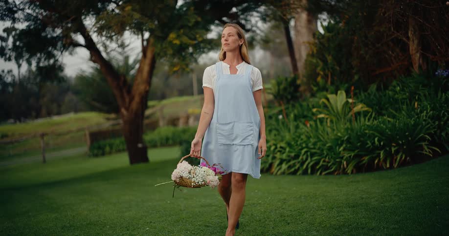 A peaceful woman in a blue apron walks through a beautiful garden in slow motion carrying a basket of fresh flowers
