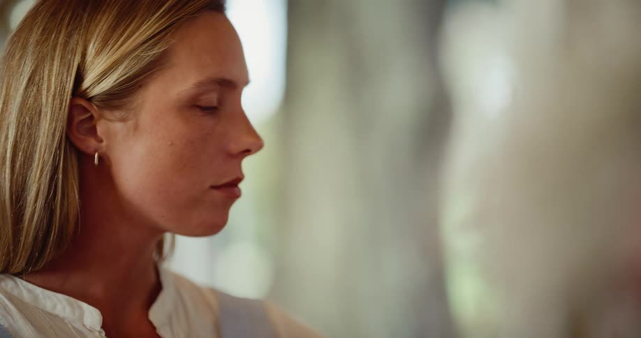 A thoughtful blonde woman with a pensive expression rests her chin on her hand while sitting indoors
